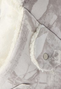 Κοντινή λήψη μπεζ μπουφάν με σχέδια, εσωτερική επένδυση από λευκή φλις και κουμπί πατιλέτας στην τσέπη.