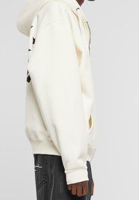Sudadera blanca con cremallera y capucha, cuenta con bolsillos tipo canguro y estampados gráficos negros en las mangas. Hecha de una tela suave con puños de canalé.