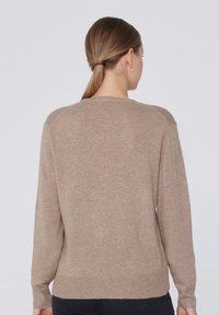 Femme aux cheveux châtain clair attachés en queue de cheval basse, portant un pull à manches longues beige et un pantalon foncé, vue de dos sur un fond clair uni.