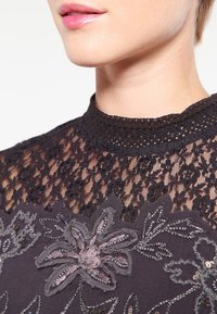 Top de encaje negro con cuello alto, que presenta intrincados bordados florales en tonos de gris y rosa. Tejido texturizado con patrones detallados.