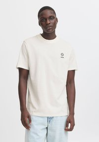 Ljusbeige bomull t-shirt med korta ärmar, rund halslinje och svart solgrafik. Bärs med ljusblå denimjeans.