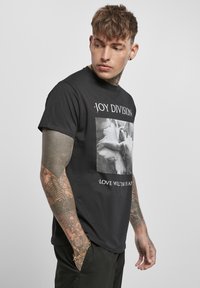 T-shirt en coton noir avec un graphique central de mains avec des ailes d'ange, texte superposé indiquant "JOY DIVISION" et "LOVE WILL TEAR US APART."