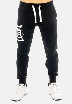 Personne portant un pantalon de survêtement noir avec l'inscription blanche "Lonsdale 1947" sur la jambe gauche et des baskets blanches avec des semelles noires, debout les pieds écartés.