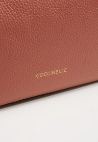 Gros plan sur un cuir rose poudré texturé avec le lettrage doré de la marque "COCCINELLE" sur un fond blanc.