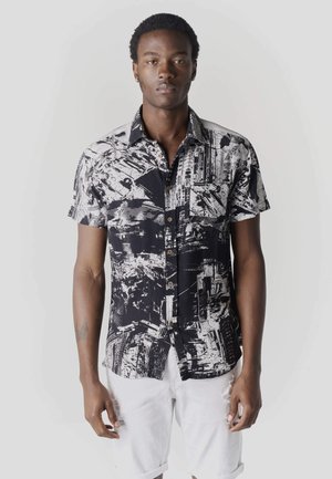 Joven con una camisa de botones de manga corta con un patrón abstracto en blanco y negro y pantalones cortos blancos, de pie frente a un fondo liso.