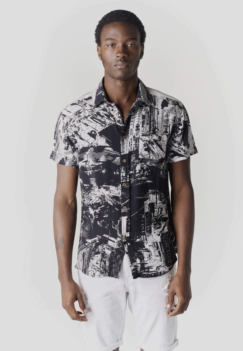 Jeune homme portant une chemise à manches courtes à boutons avec un motif abstrait noir et blanc, et un short blanc, debout devant un fond uni.