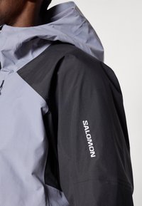 Close-up de uma pessoa a usar uma jaqueta Salomon preta e cinza com capuz, mostrando o logotipo na manga superior.