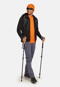 Wandelaar in een zwarte gevoerde jas, oranje shirt, grijze broek, zwarte wandelschoenen, met trekstokken en een oranje muts.