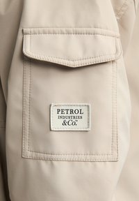 Beige lomme på et stof med et rektangulært hvidt mærkat, hvor der står "PETROL INDUSTRIES & CO." trykt i sort tekst. Syttede kanter.