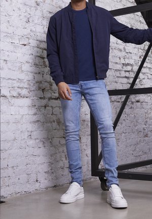 Mann in navyblauer Jacke, dunkelblauem Hemd, hellblauen Jeans und weißen Sneakers steht drinnen vor einer weißen Ziegelwand nahe schwarzen Metalltreppen.
