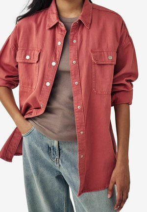 Personne portant une veste rouge ample et délavée à boutons avec poches à rabat, par-dessus une chemise marron côtelée et un jean bleu clair, une main dans la poche.