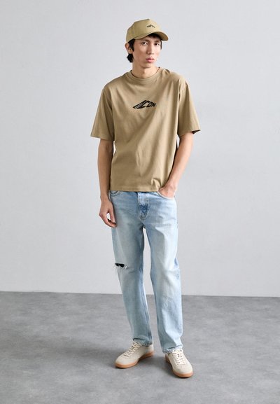 T-shirt en coton beige avec logo noir, associée à un jean déchiré bleu clair. Baskets blanches avec semelles marron clair. Casquette claire.
