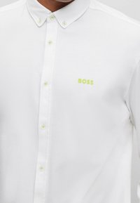 Camicia bianca con bottoni in morbido tessuto, caratterizzata da bottoni verde lime e logo sul lato sinistro del petto. Design con colletto e texture liscia.