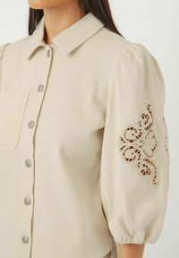 Beige skjorte med krave og sølvknapper samt en frontlomme. Ærmerne er puffede med detaljeret blondebroderi.