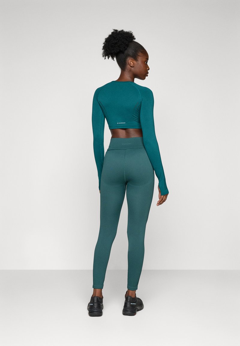 Top corto de manga larga en color verde azulado con diseño texturizado, combinado con leggings de talle alto a juego; ambos hechos de una tela elástica, usados con zapatos negros.
