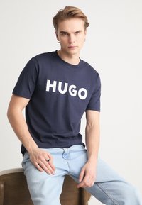 Tricou din bumbac de culoare bleumarin, cu un logo mare alb "HUGO", guler rotund, mâneci scurte și o croială lejeră. Purtați-l cu blugi deschiși.
