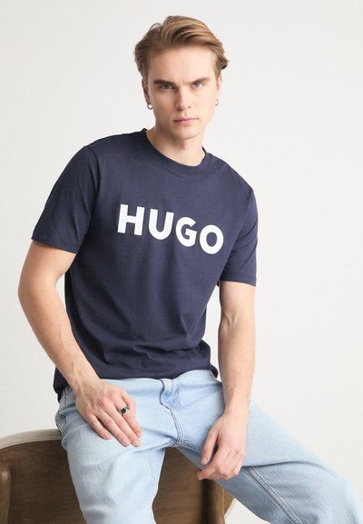Тъмносин памучен t-shirt с голям бял логотип "HUGO", с кръгло деколте, къси ръкави и свободно прилягане. С комбиниран с леки дънки.