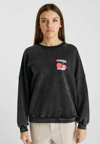 Sweat-shirt noir en tissu doux et texturé. Il présente un graphisme rose et vert avec l'inscription "AMOUREUX DES CÉRÉALES" accompagnée d'illustrations de cerises.