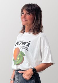 T-shirt bianco oversize con una grafica di frutti arancioni e kiwi. La scritta recita "Kiwi, sto aspettando la tua progenie." Pantaloni grigi a vita alta.