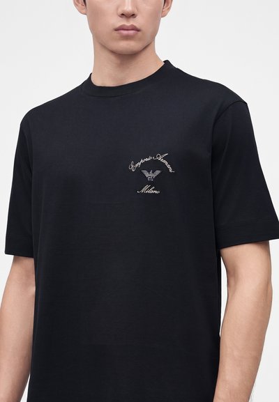 Νέος άντρας που φοράει μαύρο μπλουζάκι Emporio Armani με λευκό κεντημένο λογότυπο και κείμενο στην αριστερή πλευρά του στήθους.