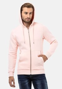 Lichtroze zip-up hoodie van zachte stof, met een capuchon, twee voorzakken en ribgebreide manchetten. Model draagt het met jeans.
