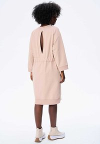 Helles rosa Sweatshirtkleid mit einem Cutout im Rücken, elastischem Taillenzug und langen Ärmeln. Kombiniert mit weißen Stiefeletten. Glatte Stofftextur.