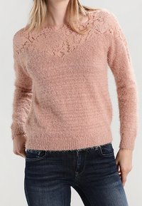 Helle rosa Sweatshirt mit einer Spitzenpasse, weichem, fluffigematerial, langen Ärmeln und gerippten Bündchen, kombiniert mit dunkelblauen Jeans.