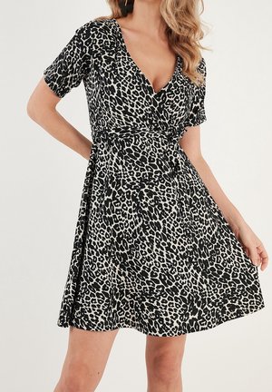 Femme portant une robe noir et blanc imprimé léopard à manches courtes et col en V, tenant la jupe de la robe d'une main.