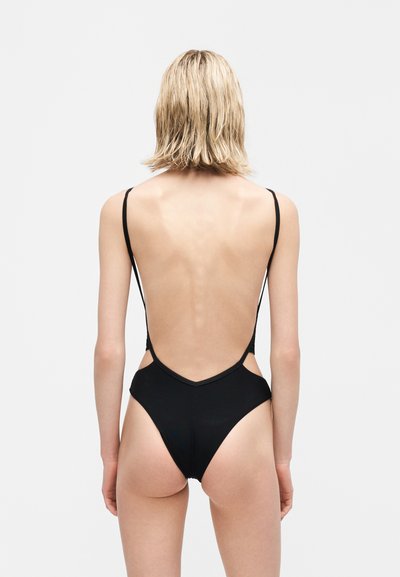 Γυναίκα με κοντά ξανθά μαλλιά φοράει μαύρο ολόσωμο μαγιό με λεπτούς ιμάντες και χαμηλή ανοιχτή πλάτη, στέκεται μπροστά σε απλό φωτεινό φόντο.