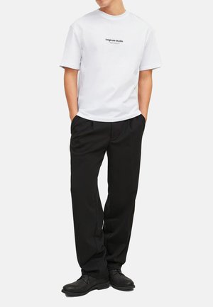 Man standing with hands in pockets wearing white "Originals Studio" t-shirt, black pants, and black shoes on a plain background.