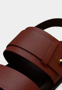 Sandales en cuir marron avec deux larges bandes, texture lisse et un accent doré. Design à bout ouvert avec un style minimaliste.