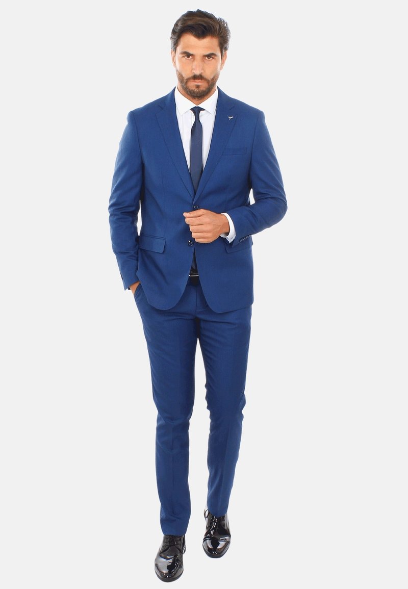 Completo blu realizzato in tessuto liscio, design slim fit, con chiusura a un bottone, camicia bianca e scarpe nere lucide.