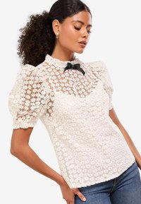 Weiße Spitzenbluse mit Puffärmeln, einem Rüschenausschnitt und einem schwarzen Schleifenakzent. Strukturierter Stoff mit transparenten Bereichen und Blumenmuster.