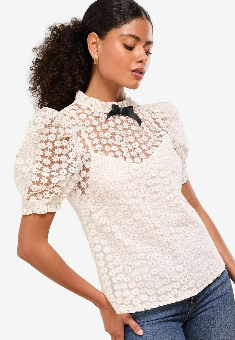 Weiße Spitzenbluse mit Puffärmeln, einem Rüschenausschnitt und einem schwarzen Schleifenakzent. Strukturierter Stoff mit transparenten Bereichen und Blumenmuster.