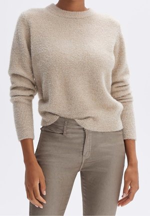 Femme portant un pull beige duveteux à manches longues, légèrement rentré dans un pantalon taupe ajusté, debout, bras détendus le long du corps.