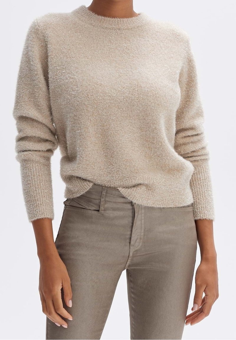 Femme portant un pull beige duveteux à manches longues, légèrement rentré dans un pantalon taupe ajusté, debout, bras détendus le long du corps.