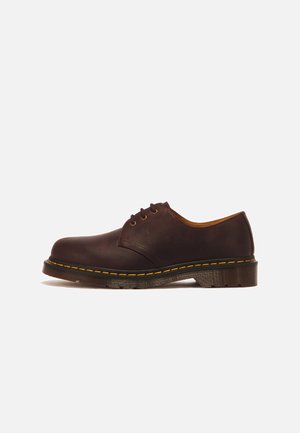 Chaussure en cuir marron foncé à lacets avec semelle épaisse et coutures jaunes sur un fond clair uni, vue de côté.