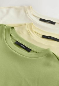 Három póló szett: egy zöld, egy halványsárga és egy krémszínű. Mindegyiknek kerek nyakkivágása van, és "NEXT" címke található a nyak hátoldalán.