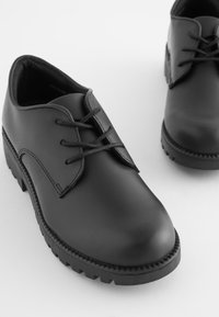 Chaussures en cuir noir avec un embout arrondi, un laçage et une semelle en caoutchouc texturée. Dessus lisse avec des coutures minimales.