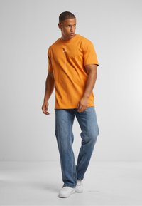 Oransje bomull t-skjorte med rund hals, korte ermer og grafisk detalj foran, kombinert med lyseblå, avslappet passform jeans og hvite joggesko.