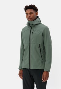 Grüne wasserdichte Softshell-Jacke mit hohem Kragen, Reißverschlusstaschen und sichtbar auf der Schulter platziertem Logo. Glatte Textur mit tailliertem Design.