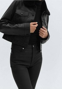 Giacca cropped in finta pelle nera con colletto morbido, chiusura a bottoni e due tasche sul petto; abbinata a jeans neri a vita alta.