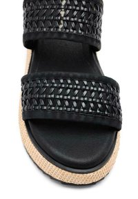 Sandalias de cuña negras con tiras trenzadas, una plantilla negra lisa y una suela texturizada de color marrón claro. Los acentos incluyen detalles trenzados en contraste.