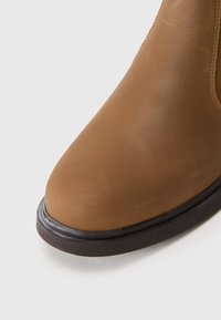 Hnědá kožená kotníková bota s kulatou špičkou, hladkým povrchem, černou gumovou podrážkou a jemným prošíváním podél strany.