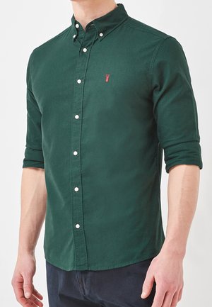 Chemise vert foncé à boutons avec un col pointu, présentant un petit logo de homard rouge sur la poitrine et des manches retroussées.