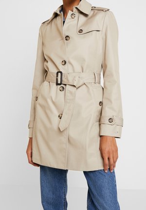 Gabardina beige con cinturón, botones, hombreras y tiras en los puños, llevada con vaqueros azules sobre un fondo liso.