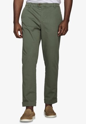 Mężczyzna w oliwkowozielonych spodniach z podwiniętymi nogawkami i brązowych wsuwanych butach, stojący na tle jednolitego jasnego tła.