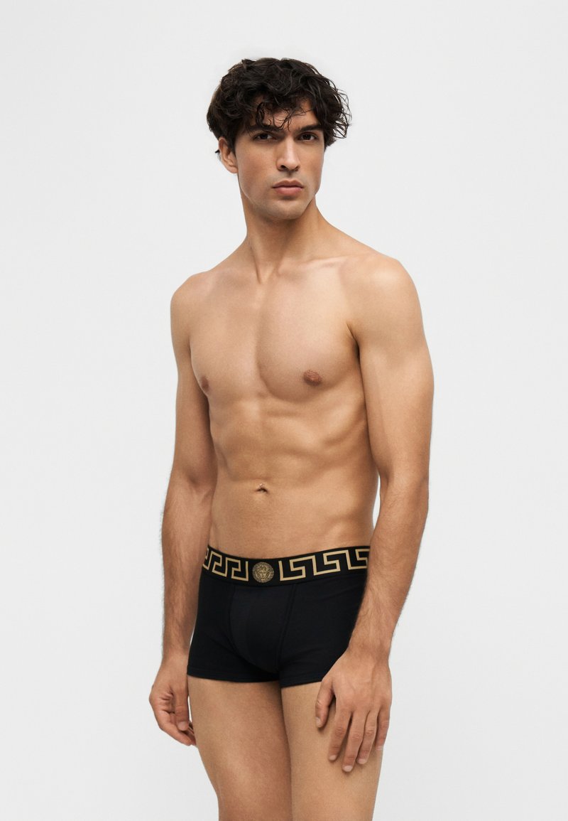 Jeune homme aux cheveux bouclés foncés portant un boxer noir avec une ceinture à motif doré, debout devant un fond clair uni.