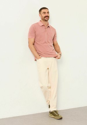 Muž s fúzmi, oblečený v ružovej polo košeli, krémových nohaviciach a zelených teniskách, opierajúci sa uvoľnene o bielu stenu, ruky v vreckách.