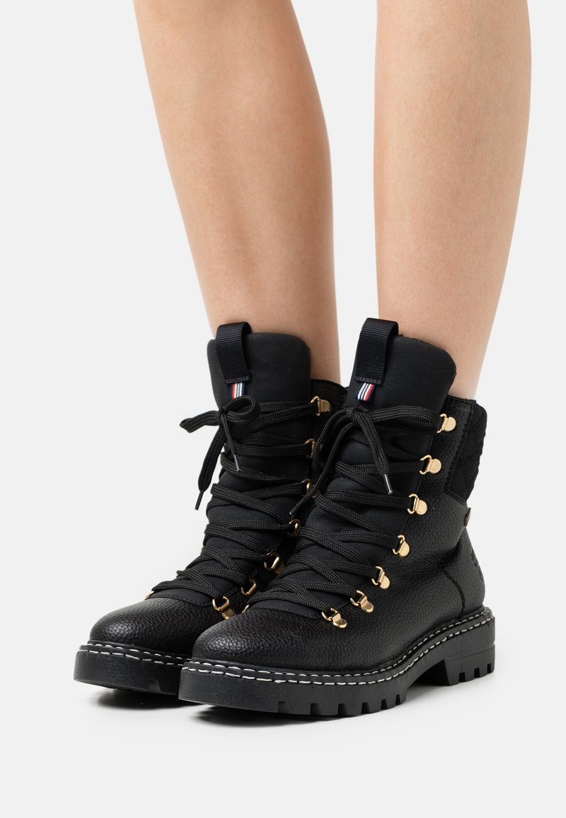Botas de senderismo de cuero negro con herrajes dorados, cordones gruesos negros y una suela robusta, con un acabado texturizado y un cuello acolchado.